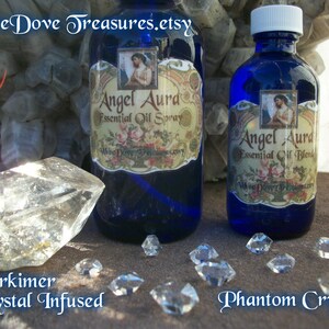 May include: Two blue glass bottles with gold labels and white text that reads "Angel Aura Essential Oil Spray" and "Angel Aura Essential Oil Blend". The bottles are sitting on a brown surface with clear quartz crystals.
