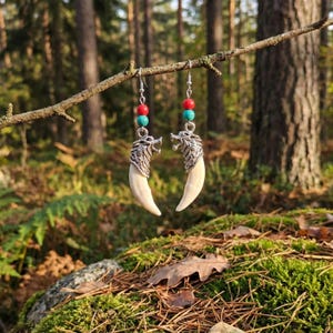 May include: Dangle earrings with a wolf head design and a large, curved, off-white tooth. The earrings have red and turquoise beads. The earrings are suspended from a branch in a forest, with a natural backdrop.