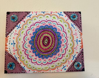 May include: A vibrant mandala artwork featuring a central circular pattern in pink, blue, green, and yellow hues. The piece is framed with a dark purple border and decorative elements. It is mounted on a white wall.