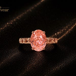 May include: A ring featuring a large, oval-cut, peach-colored gemstone set in a rose-gold band with an intricate, openwork lattice pattern. The word Monvear is printed in the upper left corner with a small four-pointed star icon above it.