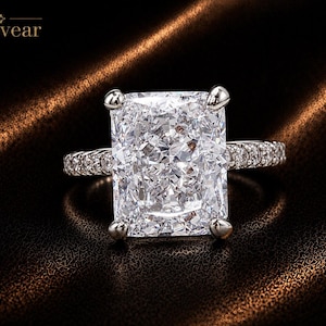 May include: A silver-toned ring featuring a large, rectangular radiant-cut center stone held by four prongs. The band is encrusted with small, round pavé-set stones along the sides. The brand name Monvear is printed in the upper left corner.