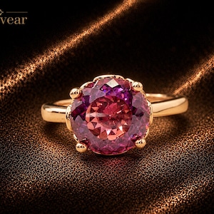 Puede incluir: Un anillo de oro con una gran piedra preciosa redonda y facetada que presenta un degradado vibrante de tonos púrpura intenso y rosa brillante. La piedra está sujeta por cuatro garras de oro en una banda pulida. El texto Monvear es visible arriba.