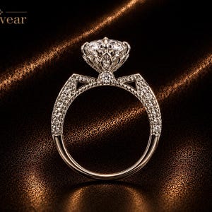Peut inclure: Une bague argentée avec une grande pierre centrale ronde et facettée, maintenue dans un sertissage décoratif en forme de pétale. L'anneau est incrusté de petites pierres scintillantes sur les côtés, et le mot Monvear est inscrit en haut à gauche.