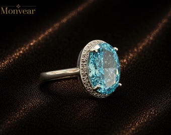 May include: A silver-toned ring featuring a large, oval-cut, light blue gemstone as the centerpiece. The central stone is surrounded by a halo of small, clear, round-cut stones set into the metal frame. The band is smooth and metallic.