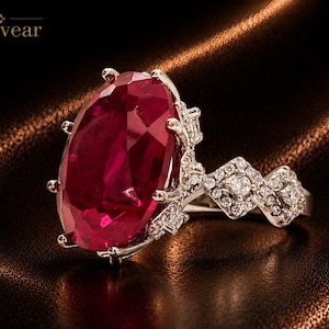 Può includere: Un anello color argento con una grande pietra preziosa rosso scuro a taglio ovale, tenuta da sei griffe. La fascia è decorata con piccole pietre chiare incastonate a pavé in un motivo geometrico. Il testo Monvear è visibile in alto a sinistra.