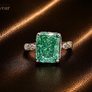 May include: A silver ring featuring a large, rectangular, radiant-cut green gemstone held by four prongs. The band is accented with small, clear, round-cut stones along the sides. The text Monvear is visible in the upper left corner of the image.