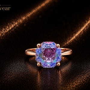 May include: A rose gold ring featuring a large, multifaceted, square-cut gemstone with a gradient of purple, blue, and pink hues. The stone is secured by four prongs on a smooth, polished band. The brand name Monvear is visible in the top left corner.