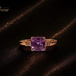 Puede incluir: Un anillo de oro con una gran piedra preciosa morada de corte cuadrado en el centro, flanqueada por dos pequeñas secciones de piedras claras engastadas en pavé en la banda. La palabra Monvear está impresa en la esquina superior izquierda con una estrella.