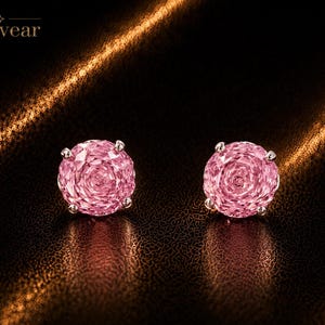Könnte beinhalten: Ein Paar runde, facettierte rosa Ohrstecker mit jeweils vier silberfarbenen Krappenfassungen. Das Wort Monvear ist in der oberen linken Ecke abgedruckt. Die Ohrringe haben einen rosenartigen Schliff, der das Licht auf der Oberfläche reflektiert.