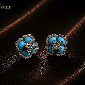 May include: A pair of stud earrings featuring a four-petaled flower design with bright turquoise stones and a central silver-toned star element encrusted with clear crystals. The edges of the petals are lined with small, sparkling clear crystals.