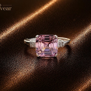 May include: A silver-toned ring featuring a large, square-cut pink gemstone as the centrepiece, held by four prongs. The band includes small, clear accent stones embedded on each side of the main setting. The brand name Monvear is visible.