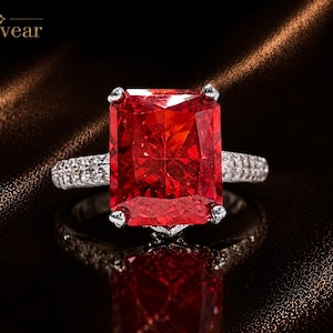 May include: A silver-toned ring featuring a large, rectangular, radiant-cut vibrant red gemstone held by four prongs. The band is encrusted with small, clear pavé-set stones. The text Monvear is visible in the upper left corner of the image.