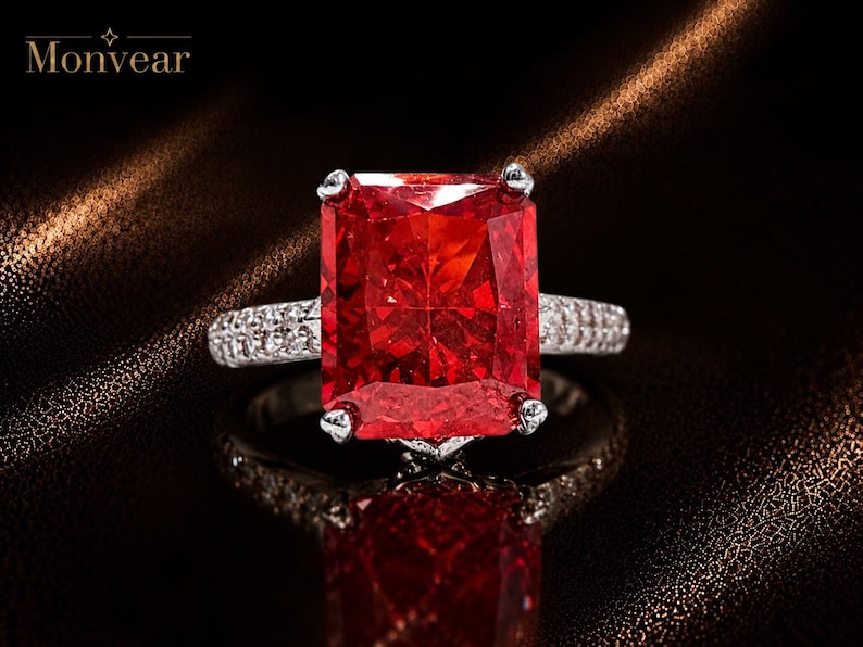 K&ouml;nnte beinhalten: Ein silberfarbener Ring mit einem gro&szlig;en, rechteckigen, im Radiant-Schliff facettierten, leuchtend roten Edelstein, der von vier Krappen gehalten wird. Die Ringschiene ist mit kleinen, klaren Pav&eacute;-Steinen besetzt. Der Text Monvear ist oben links sichtbar.