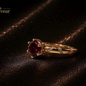 May include: A gold ring featuring a round, deep red gemstone set in a six-prong mounting, with a band accented by a series of small, clear stones set in a diagonal pattern. The word Monvear is printed in the upper left corner of the image.