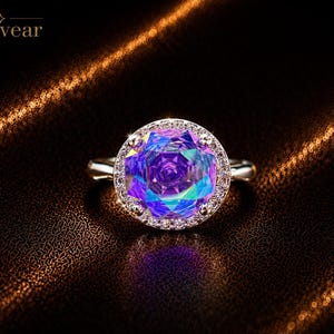 May include: A silver ring featuring a large, round, iridescent purple and blue faceted gemstone at the center, surrounded by a halo of small clear stones. The brand name Monvear is printed in the upper left corner of the image.