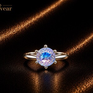 May include: A silver-toned ring featuring a round, iridescent, multi-colored gemstone set in a six-prong setting. The word Monvear is printed in the upper left corner. The gemstone displays shifting hues of blue, purple, and white light.