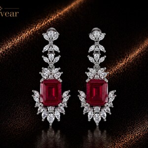 May include: A pair of drop earrings featuring a large rectangular red gemstone at the centre, surrounded by clear marquise and round cut stones in a silver-toned setting. The word Monvear is printed in the upper left corner of the image.