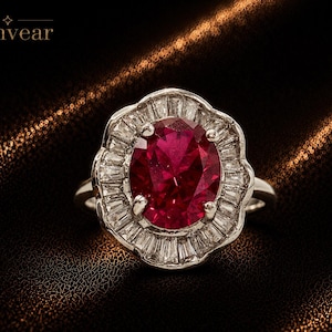 May include: A silver-toned ring featuring a large, oval-cut deep red gemstone at the center, surrounded by a halo of baguette-cut clear stones arranged in a scalloped floral pattern. The text Monvear is visible in the upper left corner.