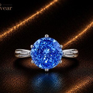 May include: A silver-toned ring featuring a large, round, brilliant-cut blue gemstone held by six prongs. The band has a split, tapered design on each side. The word Monvear is printed in the upper left corner of the image.