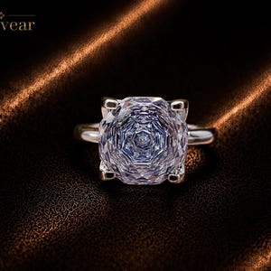 Peut inclure: Une bague argentée avec une grande pierre précieuse transparente, circulaire et à facettes, présentant un motif géométrique en taille rose. La monture comporte quatre griffes retenant la pierre. Le mot Monvear est inscrit en haut à gauche.