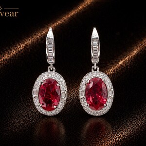 May include: A pair of silver-toned drop earrings featuring large oval-cut deep red stones surrounded by halos of small clear stones. The earring hooks are embellished with baguette-cut clear stones. The text Monvear is visible in the top left corner.