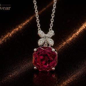 May include: A silver-toned necklace featuring a large, faceted, deep red gemstone pendant suspended from a delicate chain. Above the red stone is a small, silver-toned butterfly-shaped accent encrusted with tiny clear stones. The text Monvear is visible.