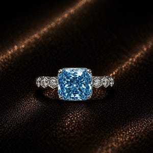 May include: A silver-toned ring featuring a large, cushion-cut, light blue center stone. The band is designed with a series of small, hexagonal settings, each containing a tiny, clear, round-cut accent stone on both sides of the center gem.