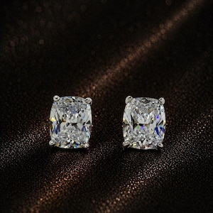 May include: A pair of stud earrings featuring cushion-cut clear stones. Each stone is held in a four-prong setting, showcasing a faceted surface that reflects light with a brilliant, colorless appearance and a symmetrical, rounded square shape.