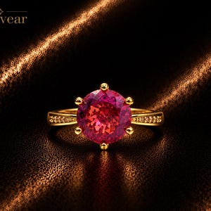 May include: A gold ring featuring a large, round, faceted pink gemstone held by six prongs. The band is accented with small, clear stones along the shoulders. The word Monvear is printed in the upper left corner of the image.
