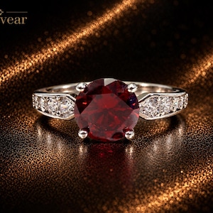 Op de afbeelding: Een zilverkleurige ring met een grote, ronde, dieprode middensteen die door vier pootjes wordt vastgehouden. De ringband is aan weerszijden van de zetting bezet met drie kleine, heldere, rond geslepen steentjes. De merknaam Monvear staat linksboven.
