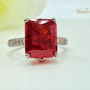 K&ouml;nnte beinhalten: Ein Ring mit einem gro&szlig;en, rechteckigen, im Radiant-Schliff geschliffenen, leuchtend roten Edelstein, der von vier Krappen gehalten wird. Die Ringschiene ist silberfarben und an den Seiten mit kleinen, klaren Pav&eacute;-Steinen besetzt. Text Monvear.