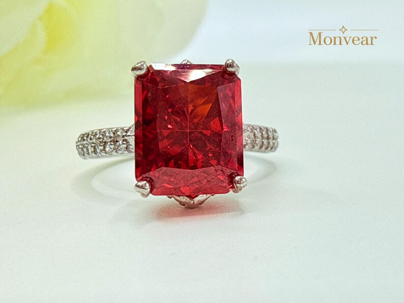K&ouml;nnte beinhalten: Ein Ring mit einem gro&szlig;en, rechteckigen, im Radiant-Schliff geschliffenen, leuchtend roten Edelstein, der von vier Krappen gehalten wird. Die Ringschiene ist silberfarben und an den Seiten mit kleinen, klaren Pav&eacute;-Steinen besetzt. Text Monvear.