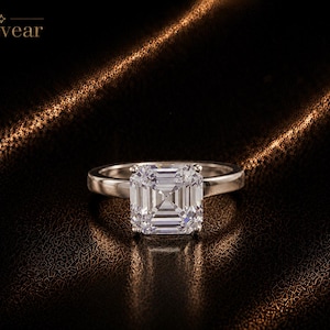 Puede incluir: Un anillo de tono plateado con una gran piedra preciosa transparente de corte cuadrado y facetas, montada en cuatro garras. La banda es lisa y pulida. La palabra Monvear está impresa en la esquina superior izquierda de la imagen.