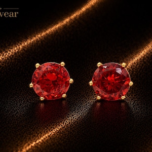 May include: A pair of round-cut red gemstone stud earrings set in gold-toned metal prongs. The earrings feature a faceted surface that reflects light, and the brand name Monvear is printed in the upper left corner of the image.
