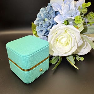 May include: A square, turquoise jewelry box with a textured surface and a gold-colored trim. The box has a small, round, gold-colored knob. A bouquet of white and blue flowers is in the background.