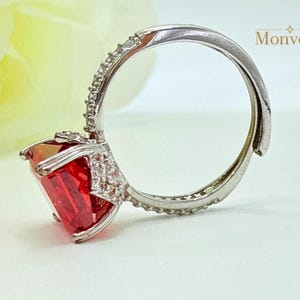 K&ouml;nnte beinhalten: Ein silberfarbener Ring mit einem gro&szlig;en, rechteckigen, leuchtend roten Edelstein in einer Krappenfassung. Die Ringschiene und die Fassung um den Stein sind mit kleinen, klaren, funkelnden Steinen besetzt. Der Text Monvear ist zu sehen.