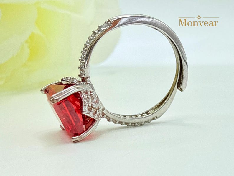 K&ouml;nnte beinhalten: Ein silberfarbener Ring mit einem gro&szlig;en, rechteckigen, leuchtend roten Edelstein in einer Krappenfassung. Die Ringschiene und die Fassung um den Stein sind mit kleinen, klaren, funkelnden Steinen besetzt. Der Text Monvear ist zu sehen.