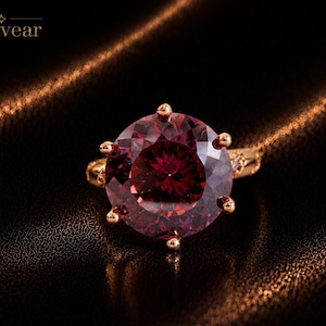 May include: A ring featuring a large, round, deep red gemstone held by six prongs on a gold-toned band. The word Monvear is printed in the upper left corner. The gemstone has a faceted surface that reflects light, and the band has a textured design.