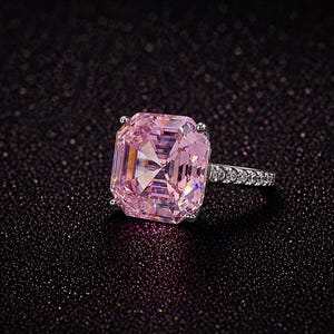 May include: A silver ring with a large, rectangular, pink gemstone. The band is set with smaller, clear stones. The ring is displayed against a dark, textured background, emphasising the sparkle of the gems.