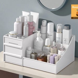 Op de afbeelding: Een witte cosmetica-organizer met twee lades en meerdere compartimenten. De organizer is gevuld met diverse huidverzorgings- en schoonheidsproducten, waaronder flessen, tubes en potjes. De producten zijn netjes gerangschikt, wat zorgt voor een overzichtelijke presentatie.