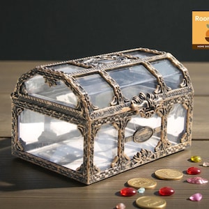 Op de afbeelding: Een decoratieve schatkist met een heldere glazen boven- en zijkanten, omlijst door sierlijk bronskleurig metaal. De kist staat op een houten oppervlak, met verspreide gouden munten en kleurrijke edelstenen.