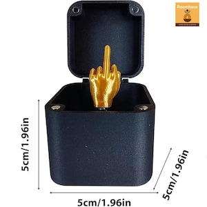 Op de afbeelding: Zwarte doos met een scharnierend deksel, met een goudkleurige hand met de middelvinger omhoog. De doos is 5 cm breed en 5 cm diep. De tekst "Roomhaus" staat in de rechterbovenhoek.