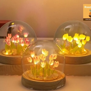 May include: Three glass dome lights featuring illuminated tulip flower arrangements. Each dome sits on a wooden base. The tulips are pink and yellow, with a warm glow. The Roomhaus logo is in the upper right corner.