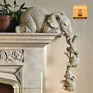 Può includere: Una scultura decorativa a forma di elefante di colore grigio chiaro. La scultura presenta un grande elefante appoggiato su una sporgenza, con tre elefanti più piccoli appesi alla sua proboscide. Un oggetto di arredamento per la casa.
