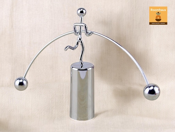 A large-scale balancing man kinetic sculpture in metal — gravity-defying equilibrium art that commands attention on any desk or shelf at a premium price.