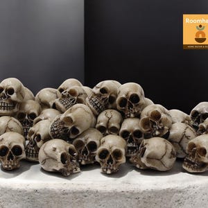 Puede incluir: Un montón de pequeñas figuras de calaveras blanquecinas. Las calaveras están apiladas unas sobre otras, con detalles visibles de las cuencas de los ojos, la cavidad nasal y los dientes. El fondo es gris oscuro.