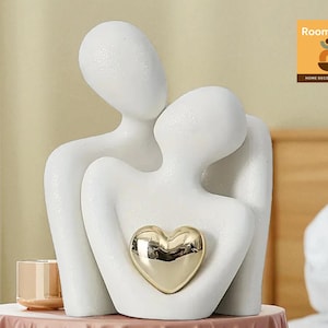 Puede incluir: Escultura de cerámica blanca que representa dos figuras abrazadas, con un corazón dorado. El diseño abstracto sugiere un tema de amor y conexión. La escultura está colocada sobre una mesa redonda con un mantel rosa. También se ve un pequeño portavelas dorado.