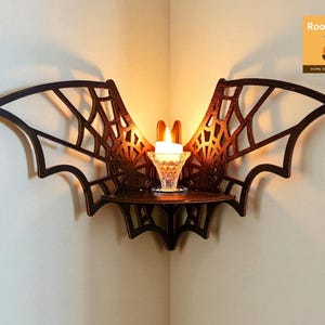 May include: A dark brown bat-shaped wall shelf with a candle holder. The shelf is made of wood and has a decorative web design. A lit candle sits in a clear glass holder on the shelf. The shelf is mounted in a corner.