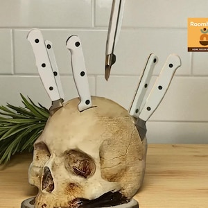May include: A decorative skull-shaped knife holder with white-handled knives. The skull is beige with dark eye sockets and a cracked texture. A sprig of rosemary is placed to the left of the skull. The knives are inserted into the top of the skull.