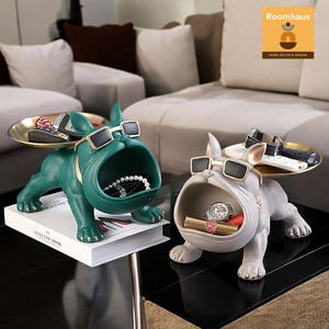 以下が含まれることがあります： ブルドッグ型の装飾トレイ2つ。1つはティール、もう1つはグレーです。どちらもサングラスをかけており、収納用に口が開いています。ティールのブルドッグは本の上にあります。トレイには、ジュエリー、時計、リモコンが入っています。